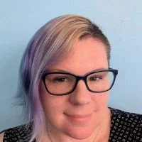 A woman with glasses and light-colored hair with hints of purple, smiling against a light blue background.