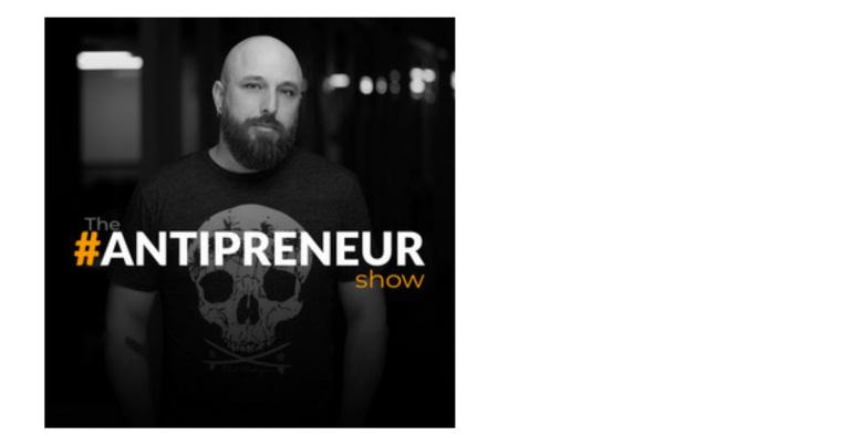Black and white photo of a man with a beard and shaved head, wearing a black T-shirt with a skull graphic, standing in a dark indoor space. Text on the image reads 'The #ANTIPRENEUR Show'.