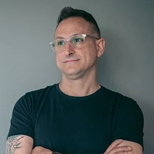 A man with short dark hair, glasses, and tattoos on his arms, wearing a dark t-shirt, standing against a plain gray background.
