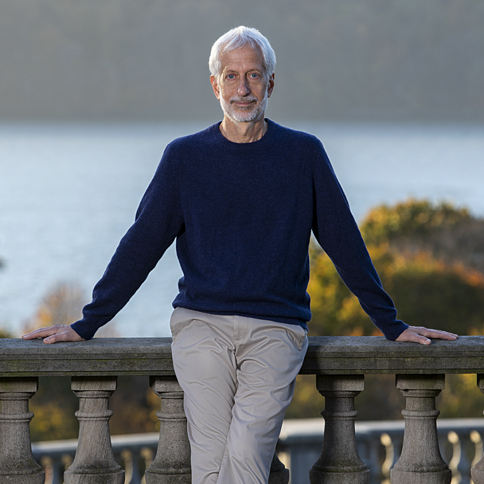 An elderly man with white hair and a beard leaning on a stone railing outdoors during daytime, with trees and water in the background.