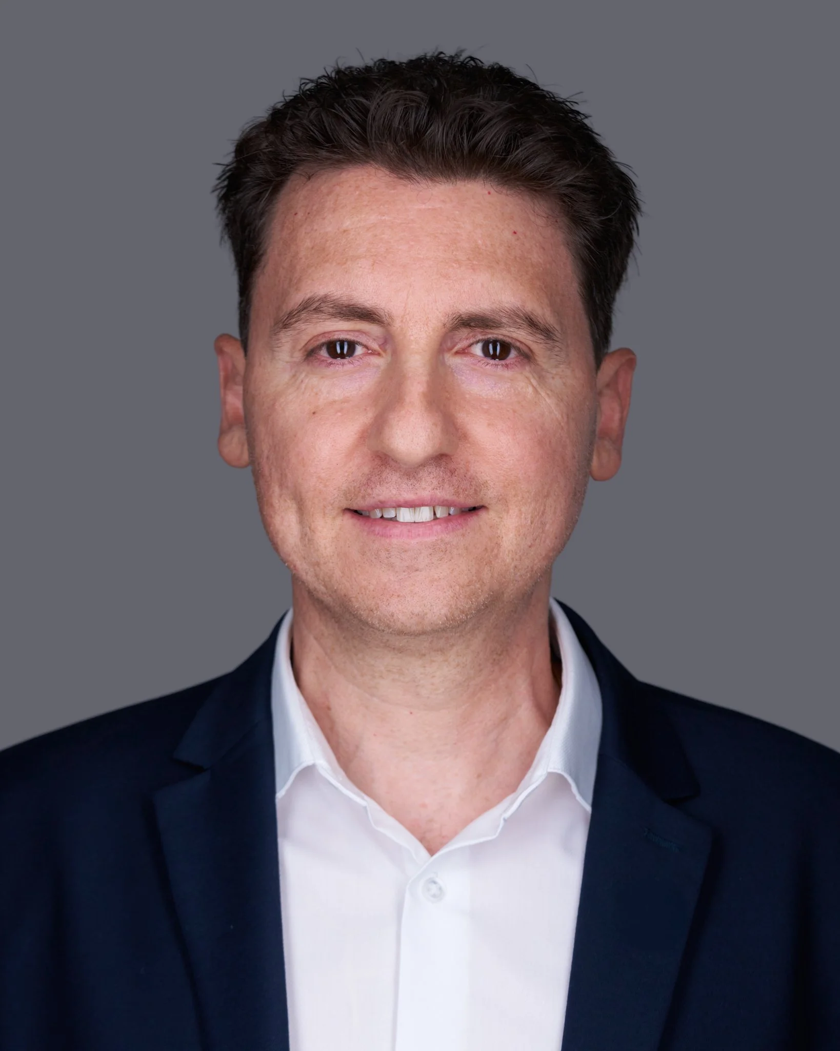 A man in a dark suit and white shirt, smiling and staring at the camera, with a gray background.