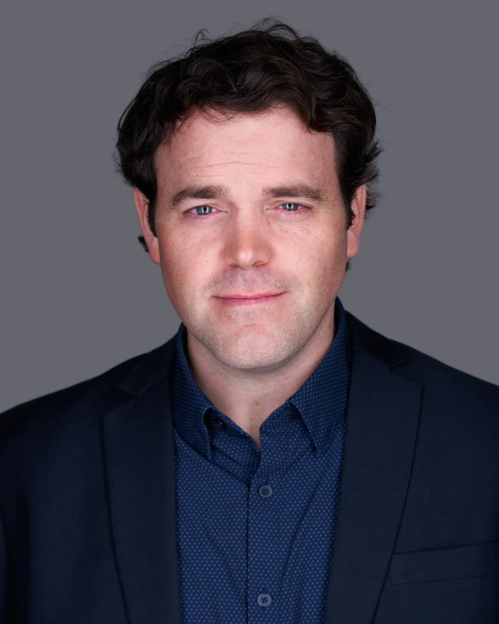 Close-up of a man with dark, wavy hair, fair skin, and blue eyes, dressed in a dark blazer and a blue, patterned shirt, against a plain grey background.