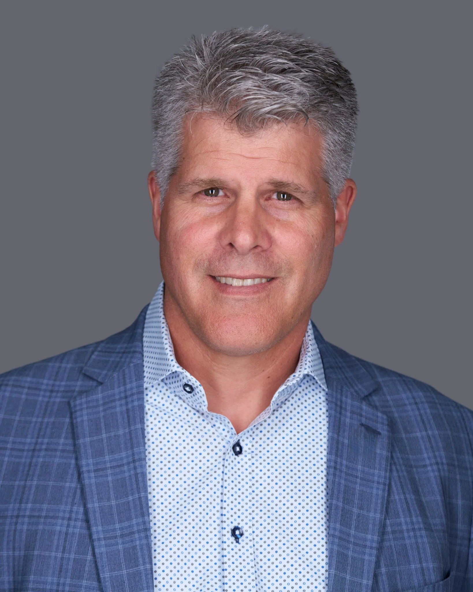 Headshot of a middle-aged man with gray hair, smiling, wearing a blue checked blazer and a white shirt with small blue polka dots, against a gray background.