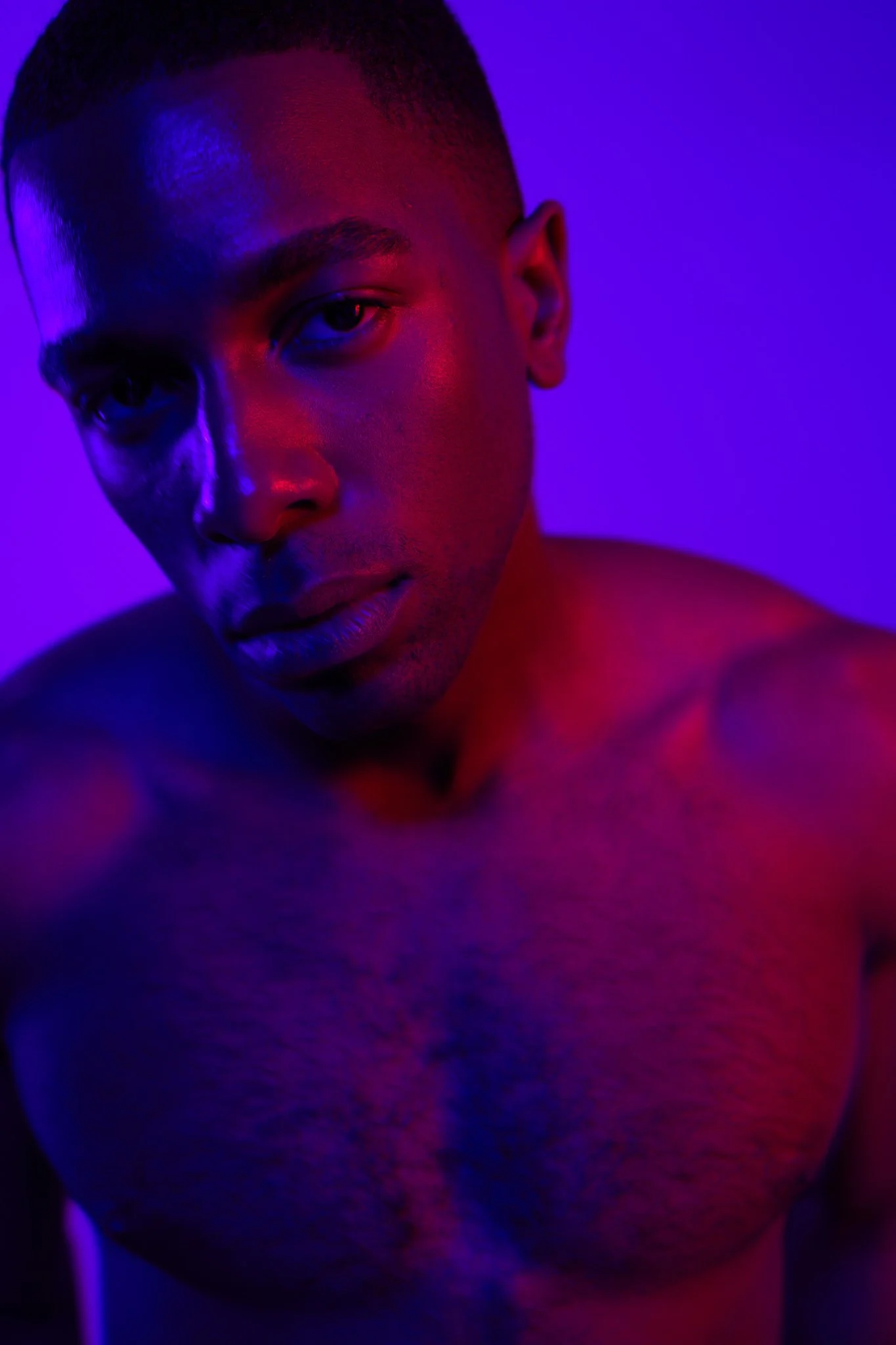Close-up portrait of a shirtless man with short hair, illuminated by purple and red lighting, against a purple background.