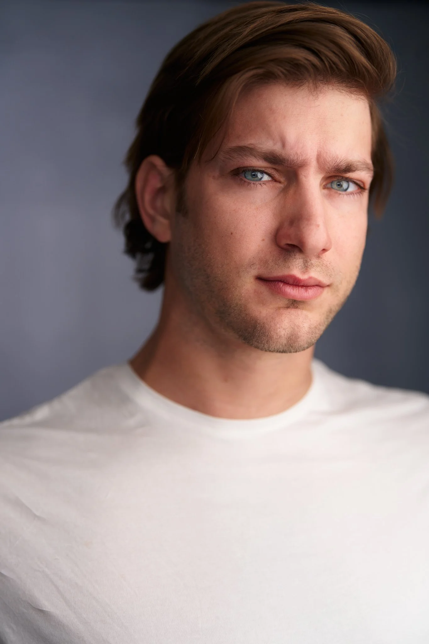 Close-up of a man with blue eyes and brown hair, wearing a white shirt, against a dark background.