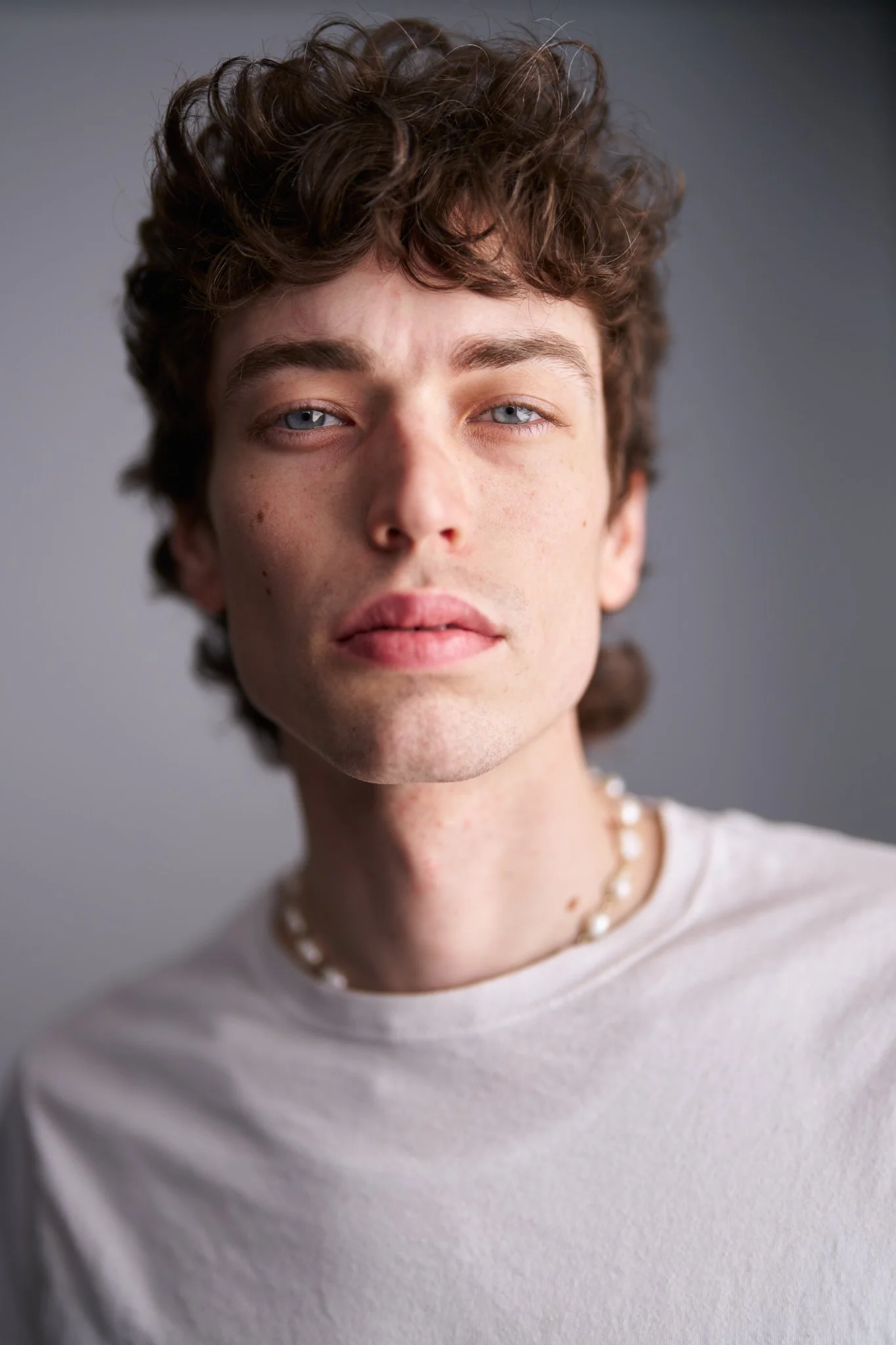 A young man with curly brown hair and blue eyes, wearing a white shirt and a pearl necklace, looking directly at the camera against a plain gray background.