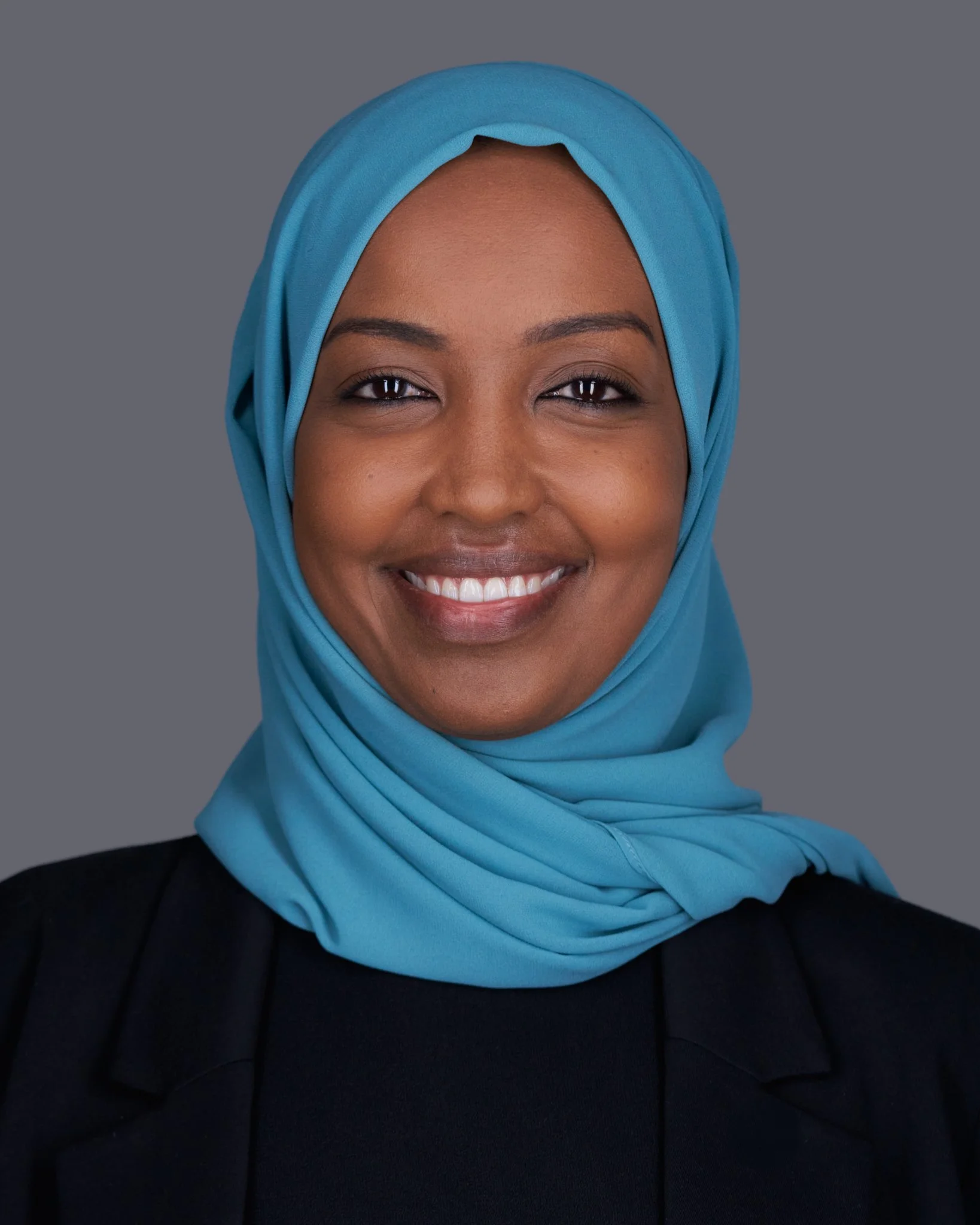 Close-up of a woman with dark skin wearing a bright blue hijab and a black top smiling against a gray background.