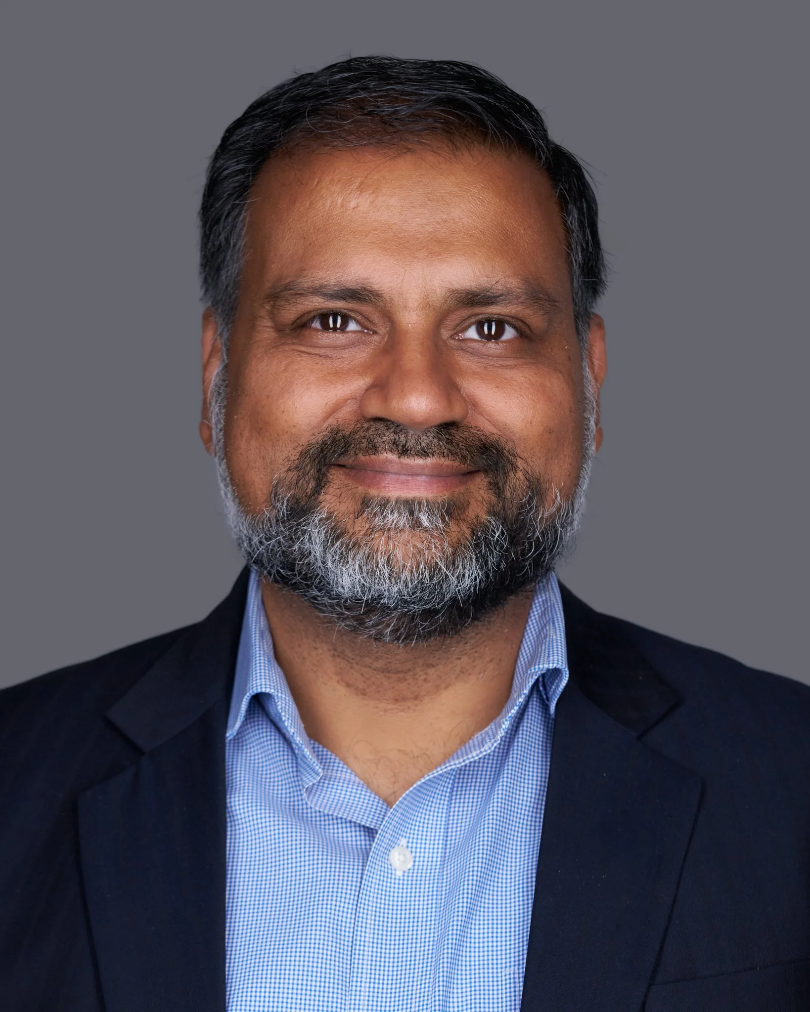 Close-up of a middle-aged man with dark hair, a beard with gray, wearing a dark blazer and a light blue button-up shirt, smiling against a gray background.