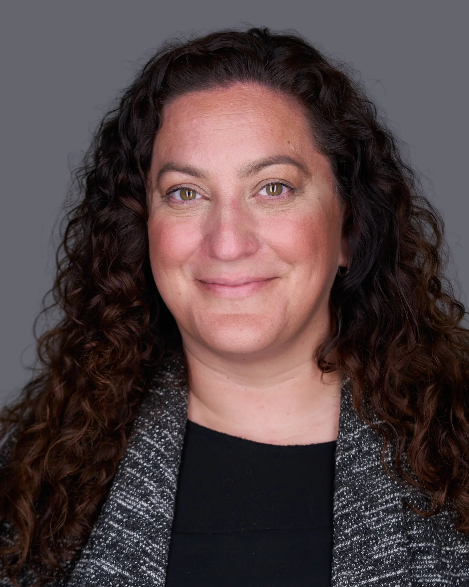 Close-up photo of a woman with long, curly dark hair, smiling softly, wearing a black blouse and a gray sparkly cardigan against a plain gray background.