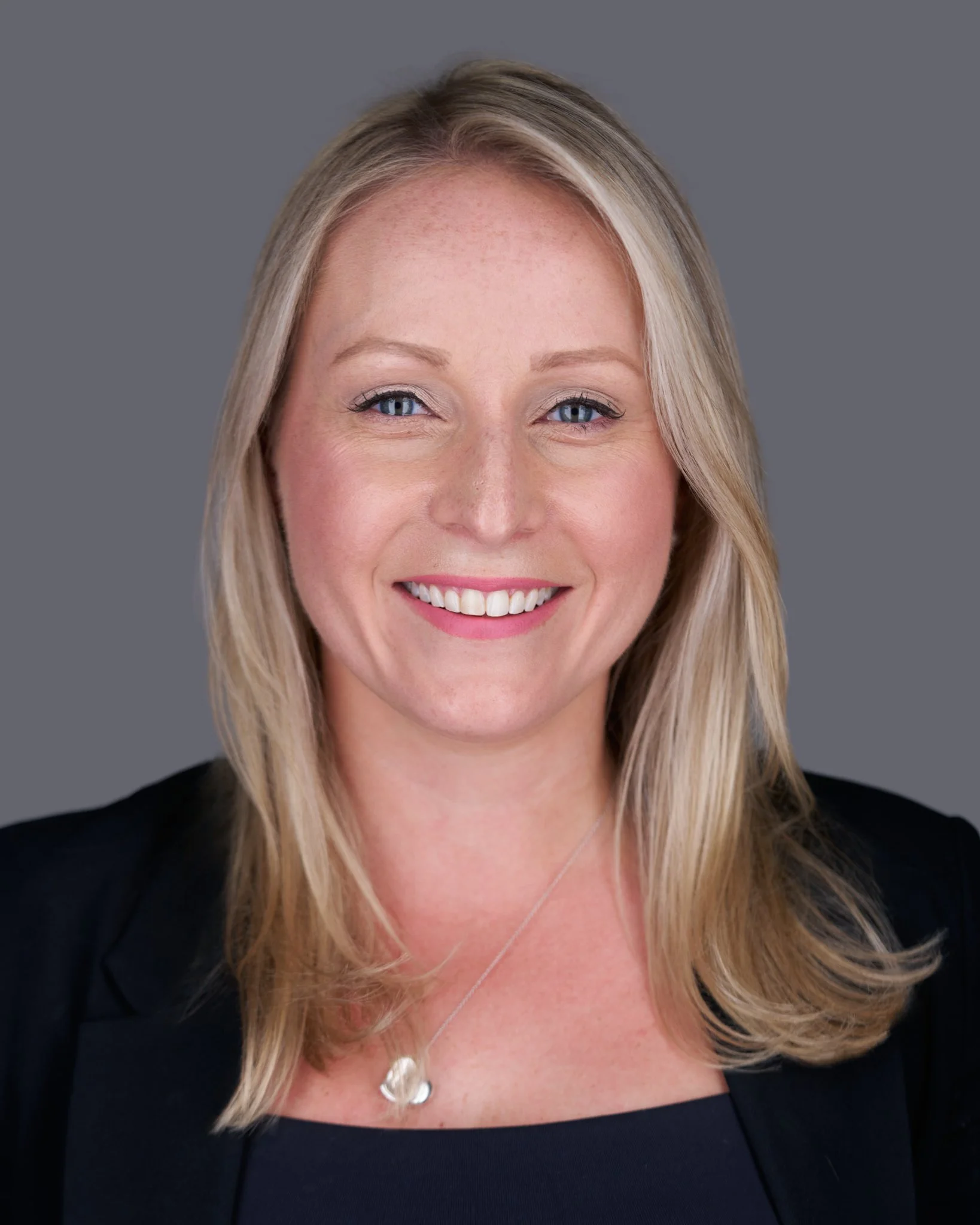 Close-up of a woman with blonde hair, smiling, wearing a black top and a silver necklace with a moon pendant, set against a grey background.