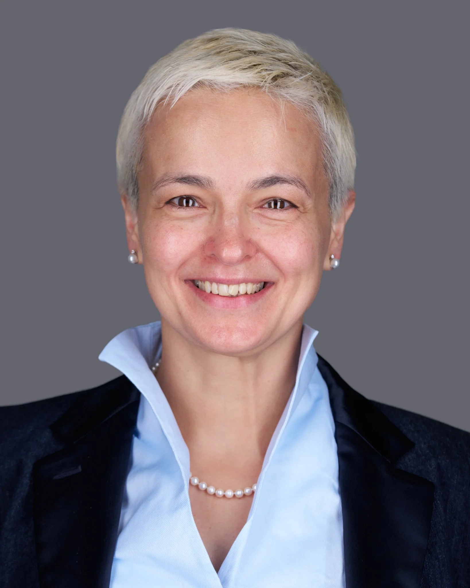 Close-up of a woman with short blond hair, smiling, wearing pearl earrings, a light blue collared shirt, and a dark blazer, against a gray background.