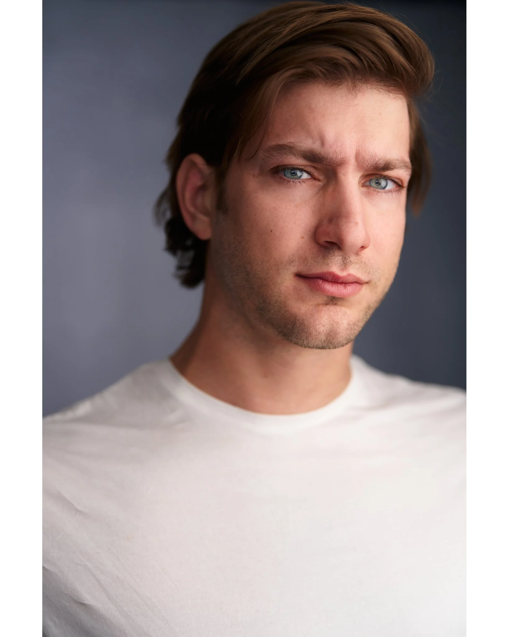 Close-up of a young man with light skin, blue eyes, brown hair, wearing a white shirt against a dark background.