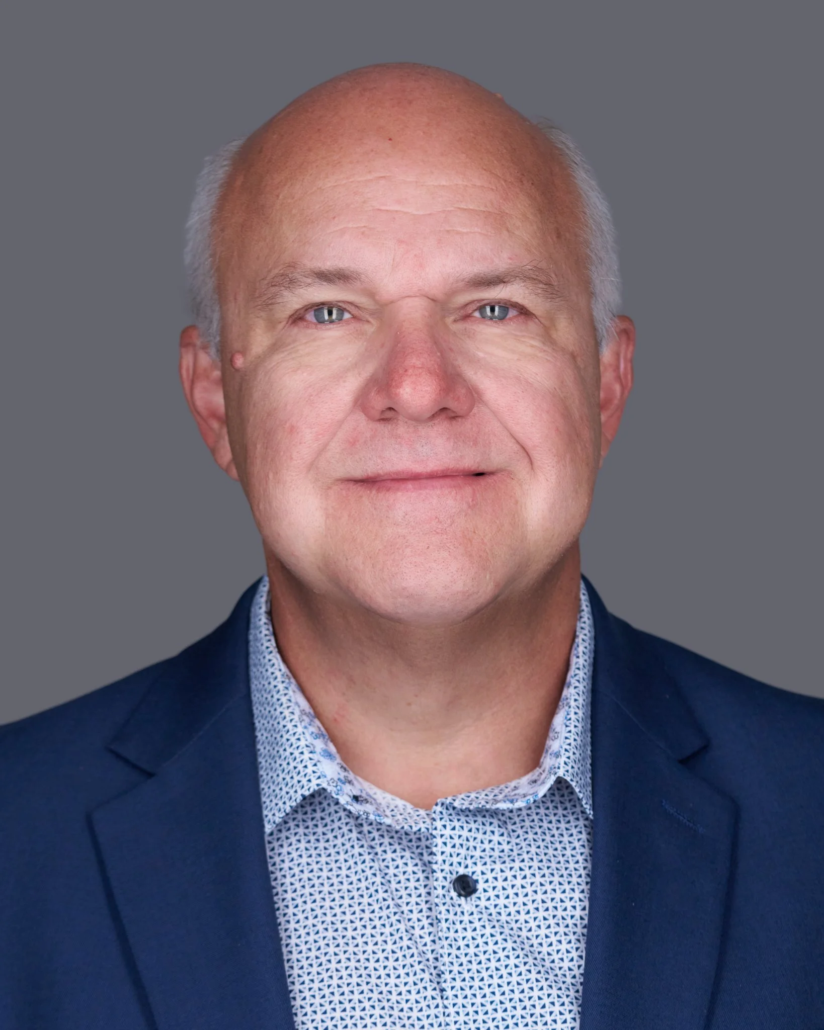 Close-up of a middle-aged man with a bald head, light skin, blue eyes, and a faint smile, wearing a navy blazer and a patterned shirt, against a plain gray background.