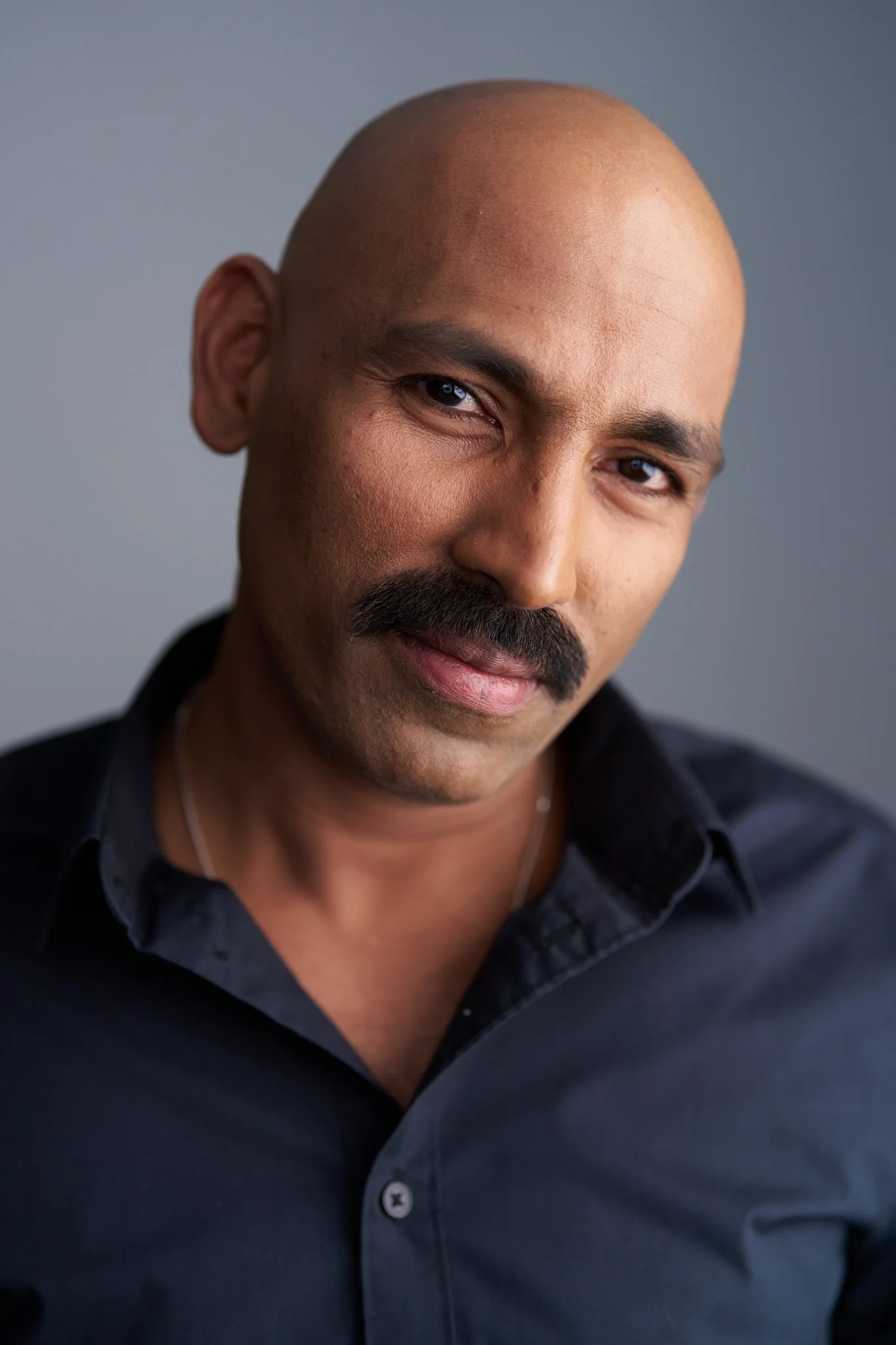 Close-up portrait of a bald man with a mustache, wearing a dark collared shirt, against a neutral background.