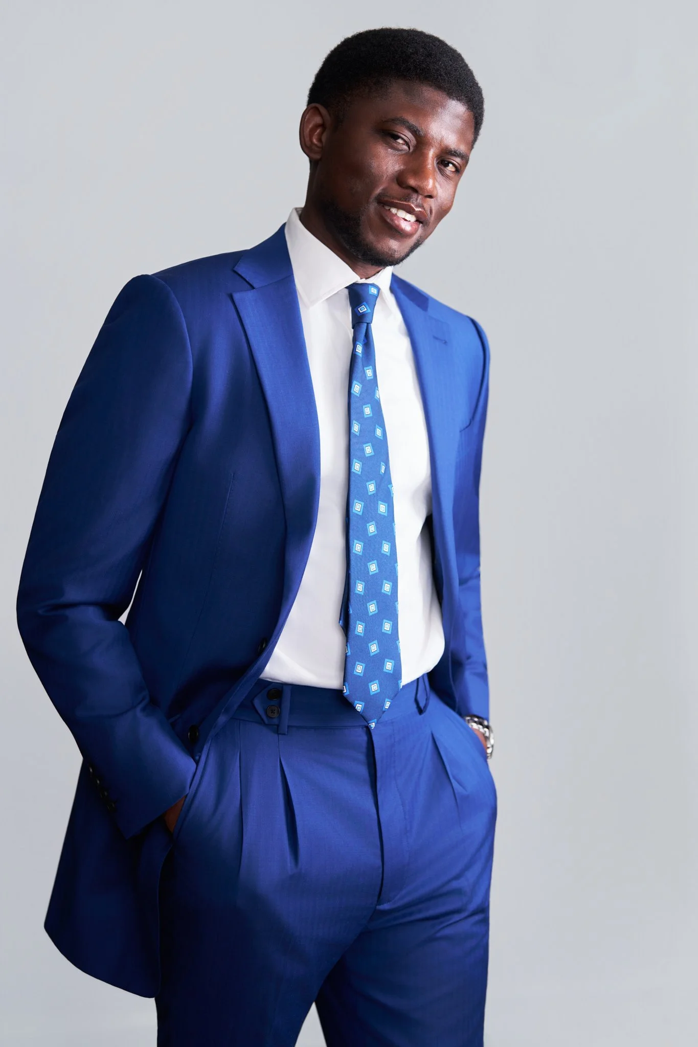A man in a blue suit with matching pants, jacket, and a patterned tie, standing with one hand in his pocket and smiling against a plain light gray background.