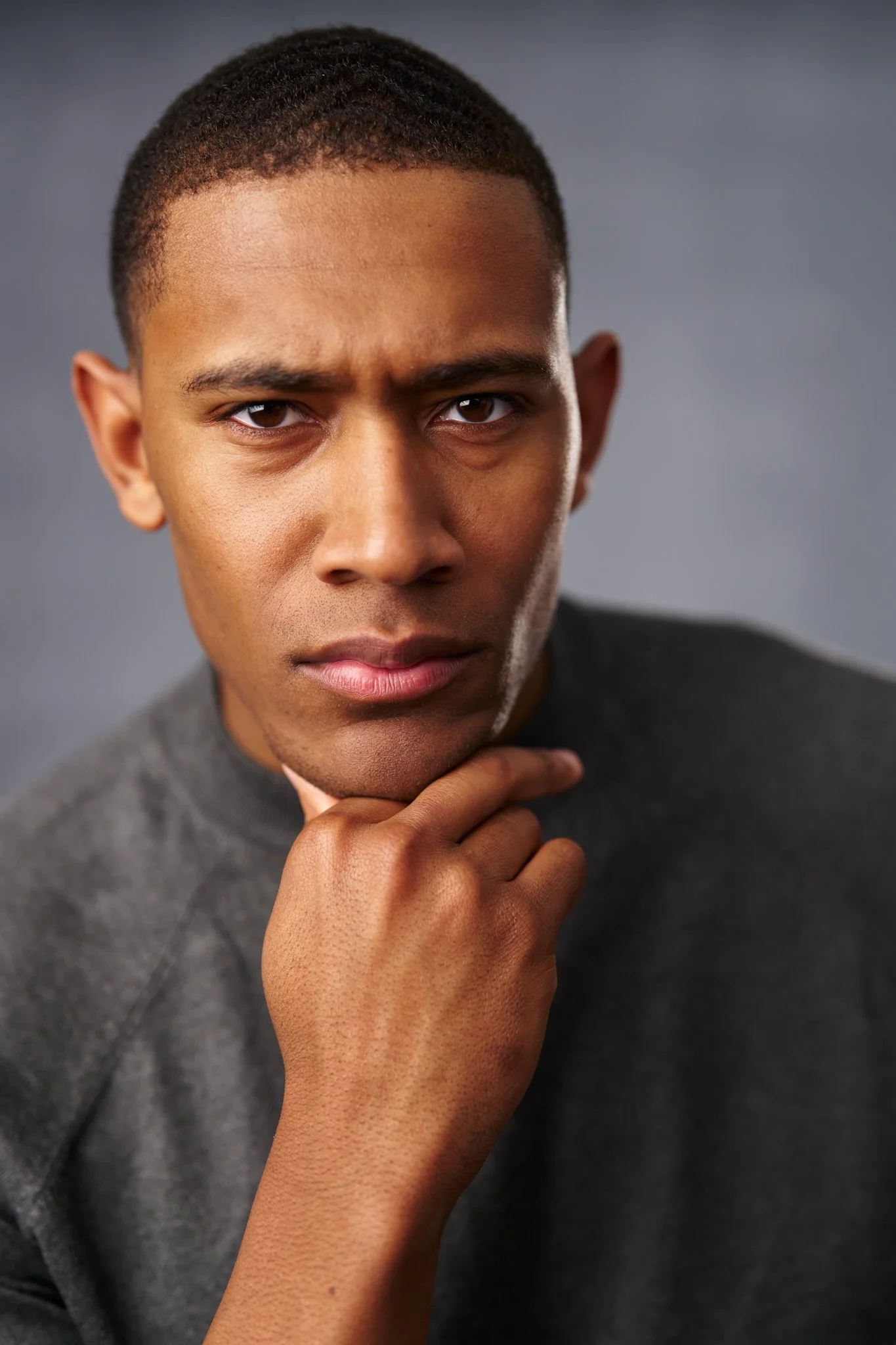 Professional headshot of a young Black man with short hair, wearing a dark gray sweater, resting his chin on his hand against a gray studio backdrop with dramatic side lighting.
