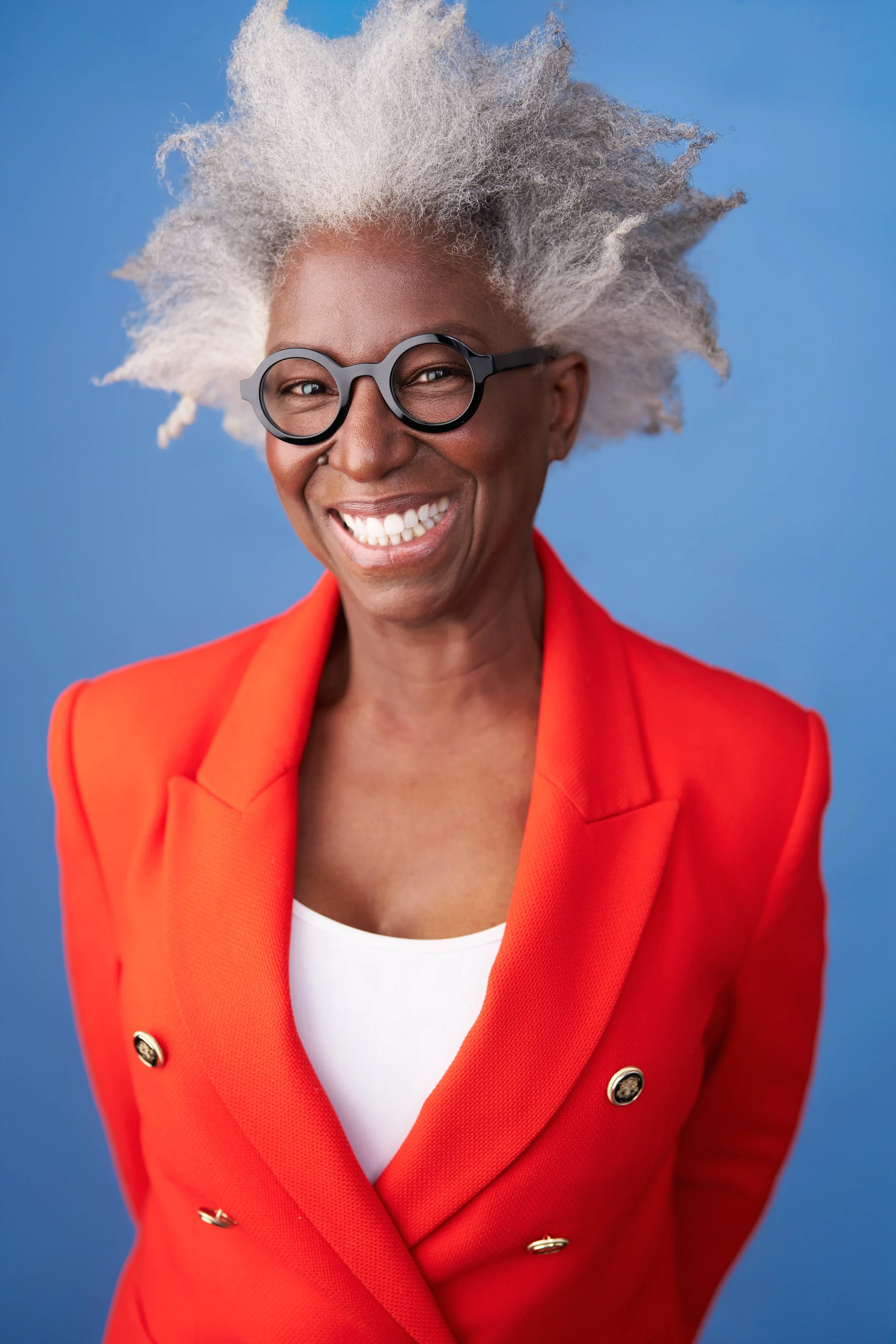 A smiling woman with gray hair and glasses wearing a bright red blazer and white top against a blue background.