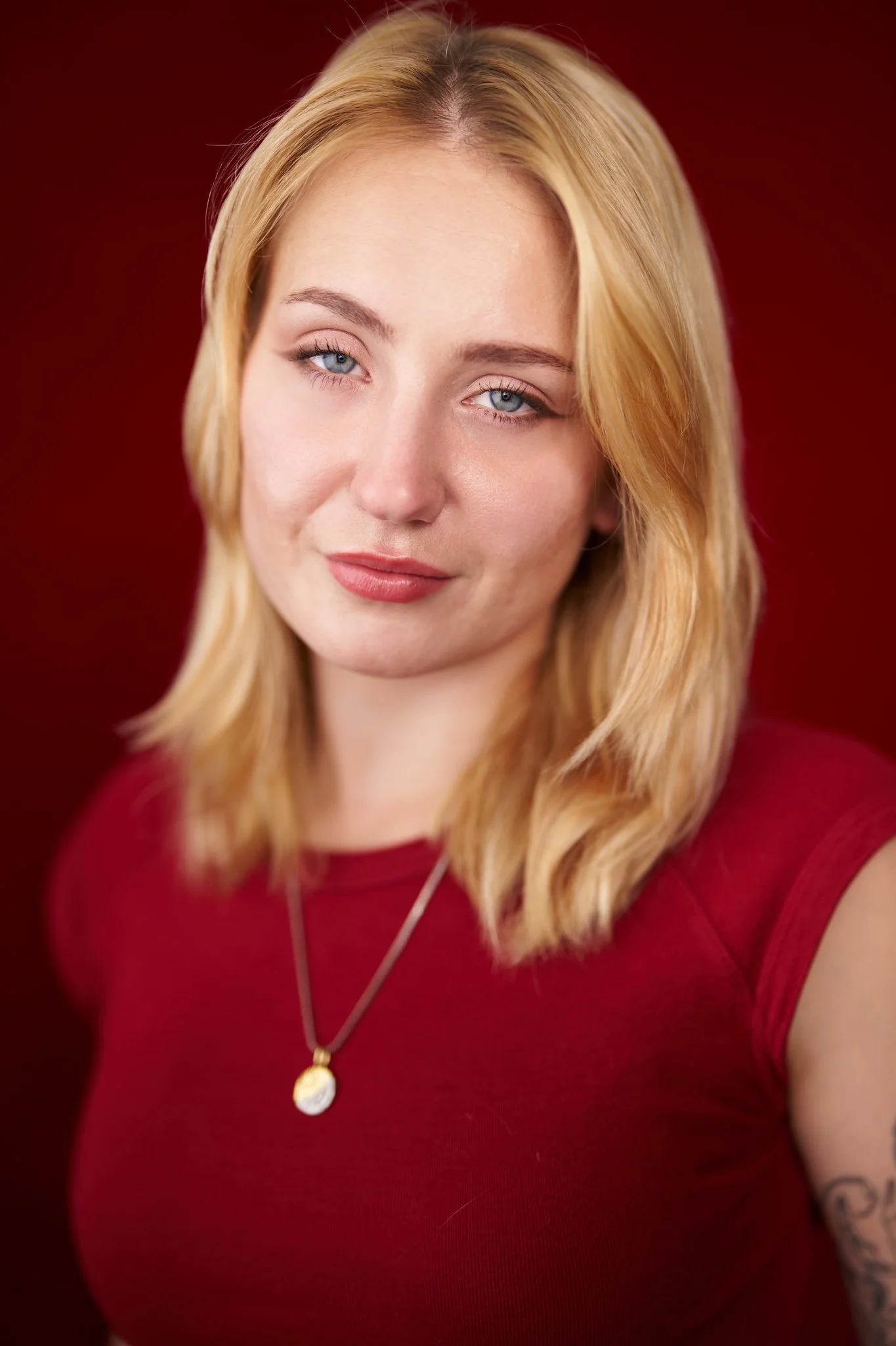 Close-up portrait of a woman with blonde hair, blue eyes, and wearing a red top and gold necklace, against a red background.