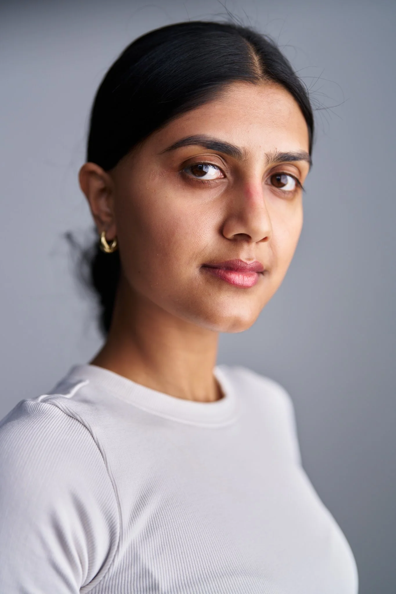 Close-up portrait of a woman with dark hair tied back, wearing a white shirt, standing against a plain gray background.