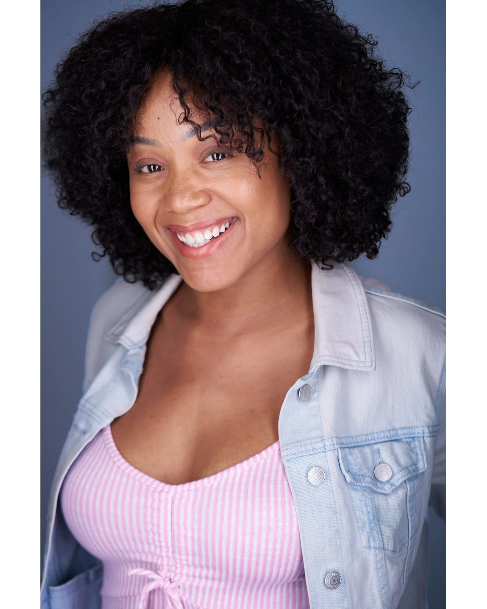 Smiling woman with curly black hair, wearing a pink and white striped top and a light blue denim jacket, against a gray background.