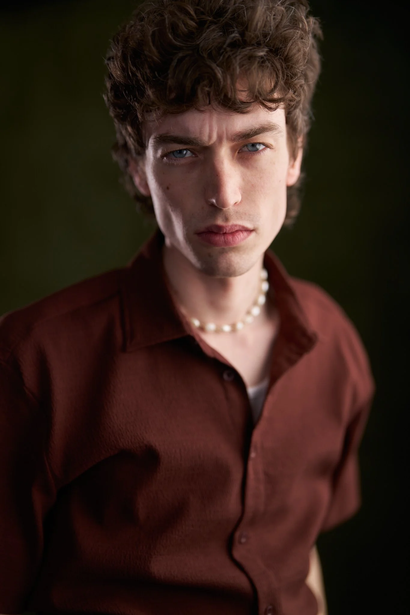 Close-up of a young man with curly brown hair, blue eyes, wearing a maroon shirt and a pearl necklace, with a dark background.