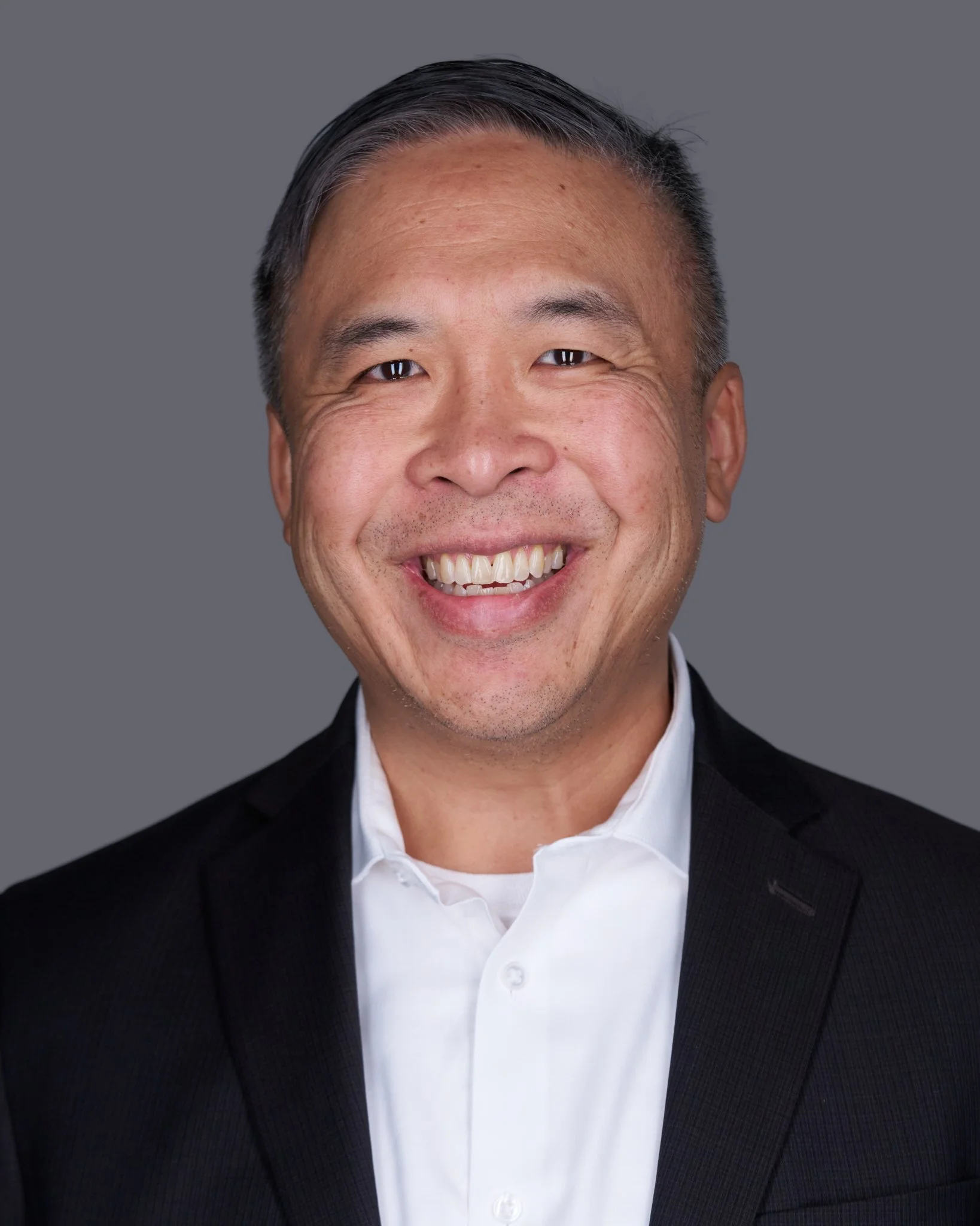 Smiling man in a black suit, white shirt, and tie against a gray background.