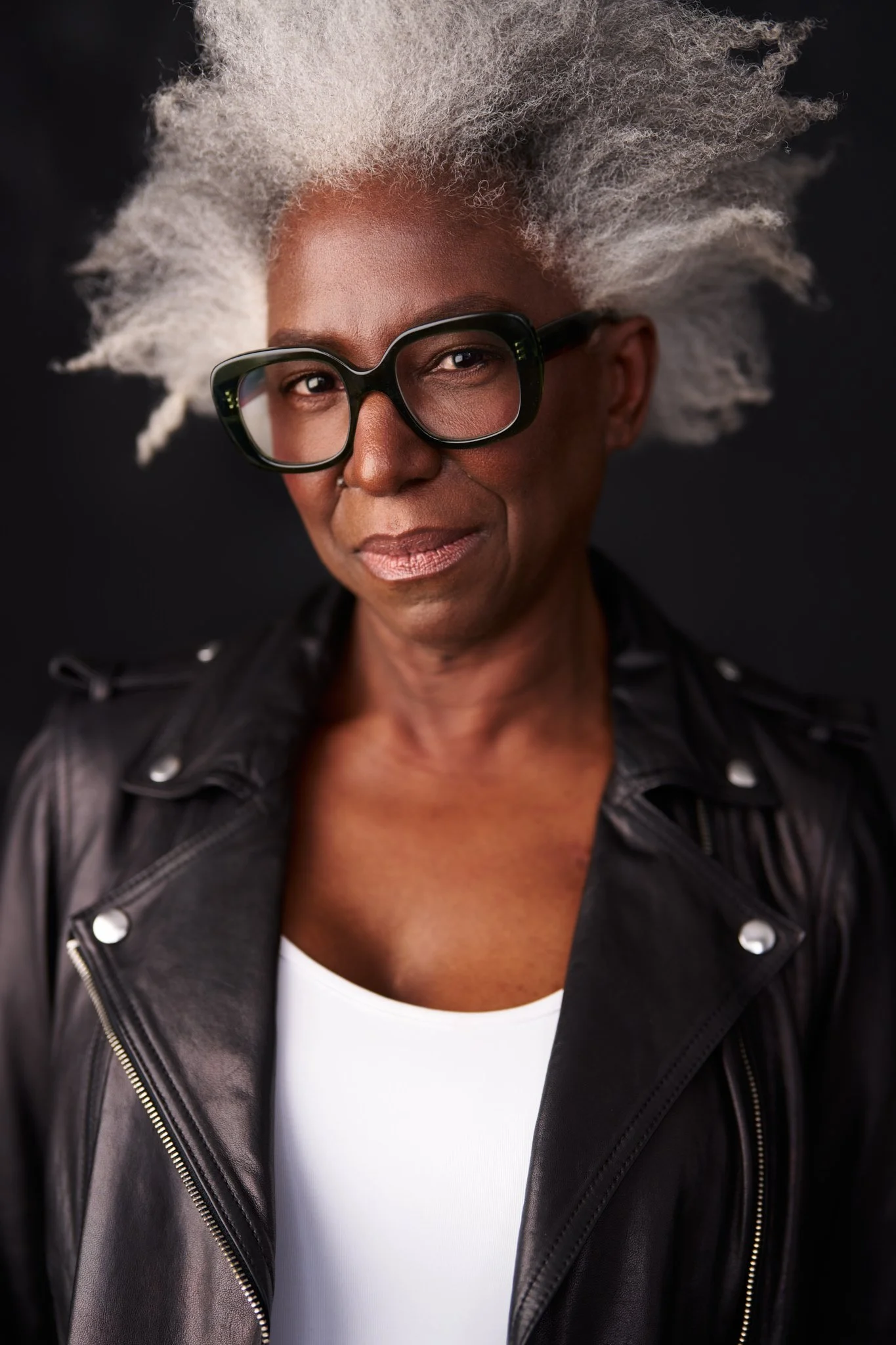 Close-up portrait of an African American woman with short gray curly hair, wearing large black glasses, a black leather jacket, and a white top, against a black background.