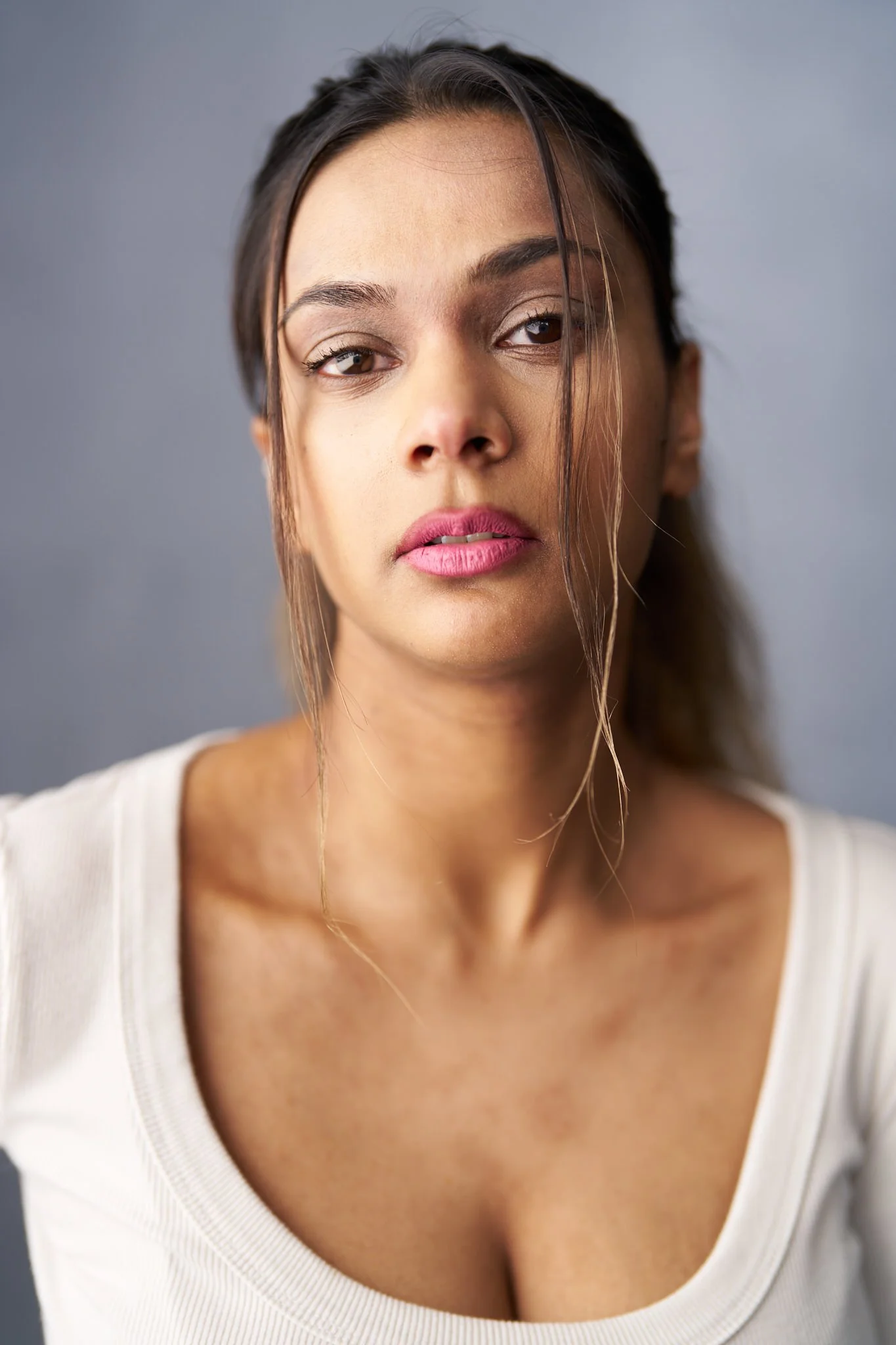 A young woman with medium brown skin and long dark hair, wearing a white top, looking into the camera with a neutral expression against a gray background.