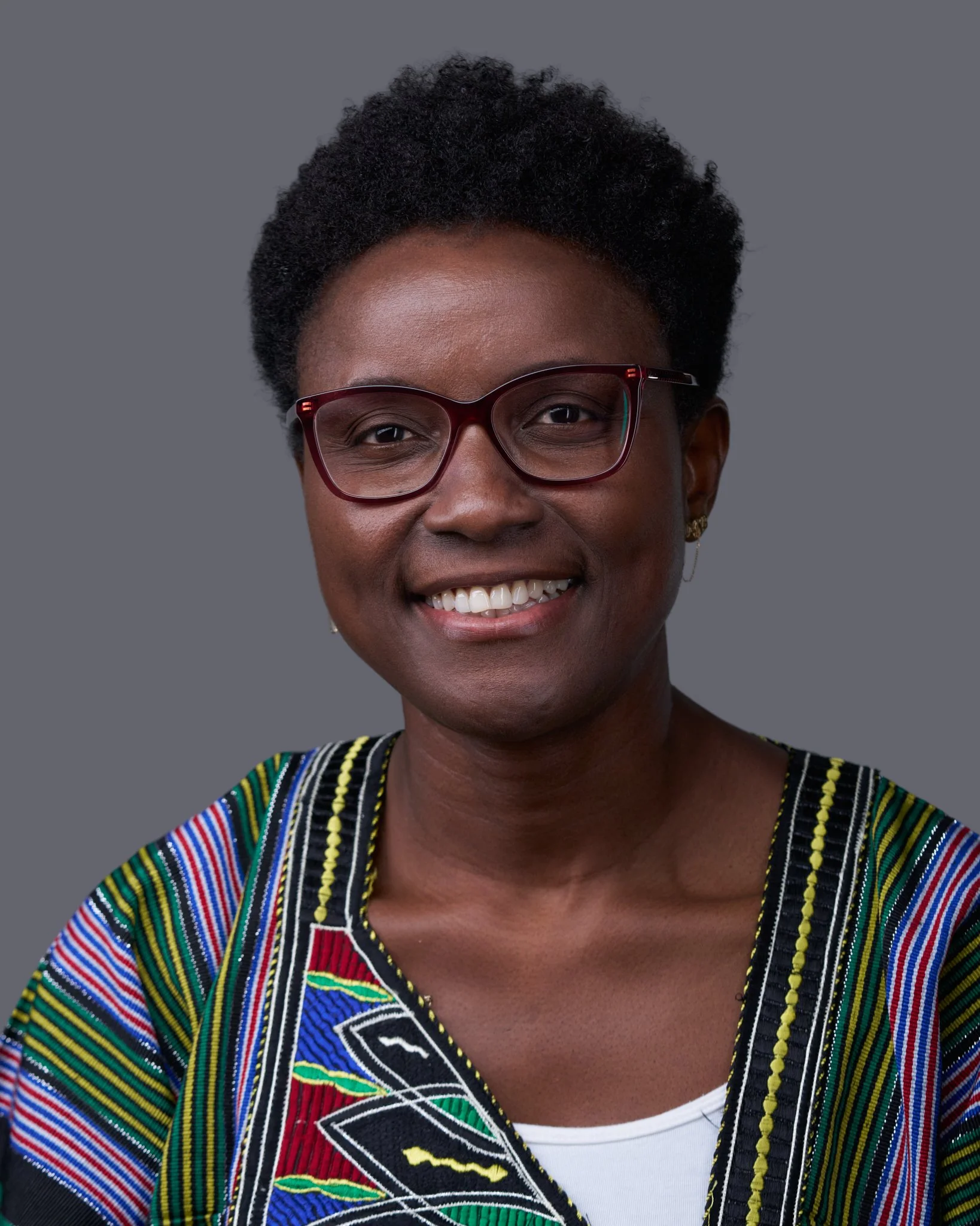 Close-up of an African American woman smiling, wearing glasses and a colorful, patterned traditional African dress with a white inner shirt.