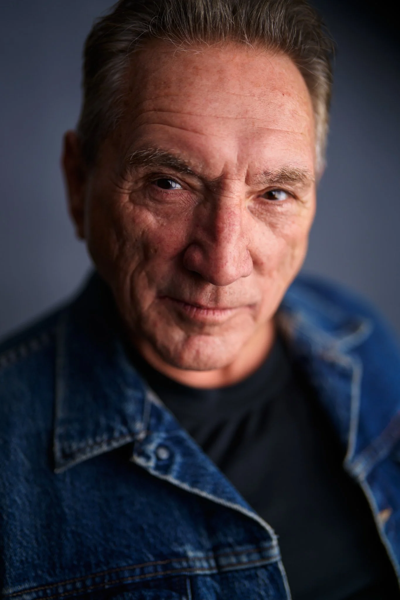 Close-up portrait of a mature man with light skin, wearing a denim jacket, looking confidently into the camera.