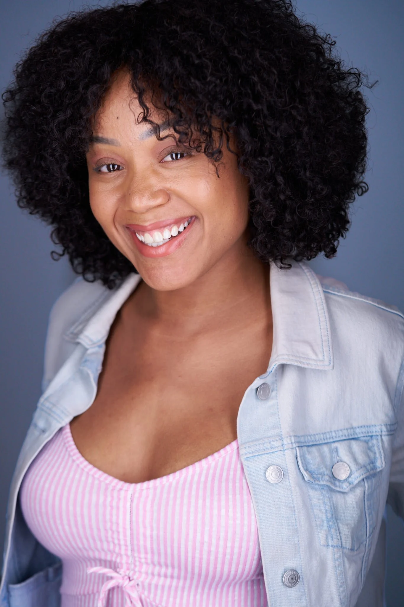 Close-up of a woman smiling, wearing a light denim jacket over a pink and white striped top, with curly dark hair and a solid dark background.