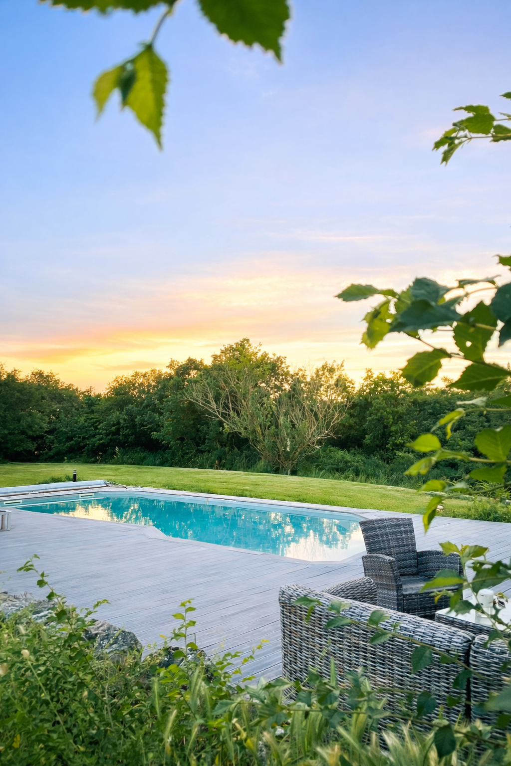 Piscine extérieure entourée de végétation avec un ciel pastel au crépuscule et des chaises en osier noir et blanc.