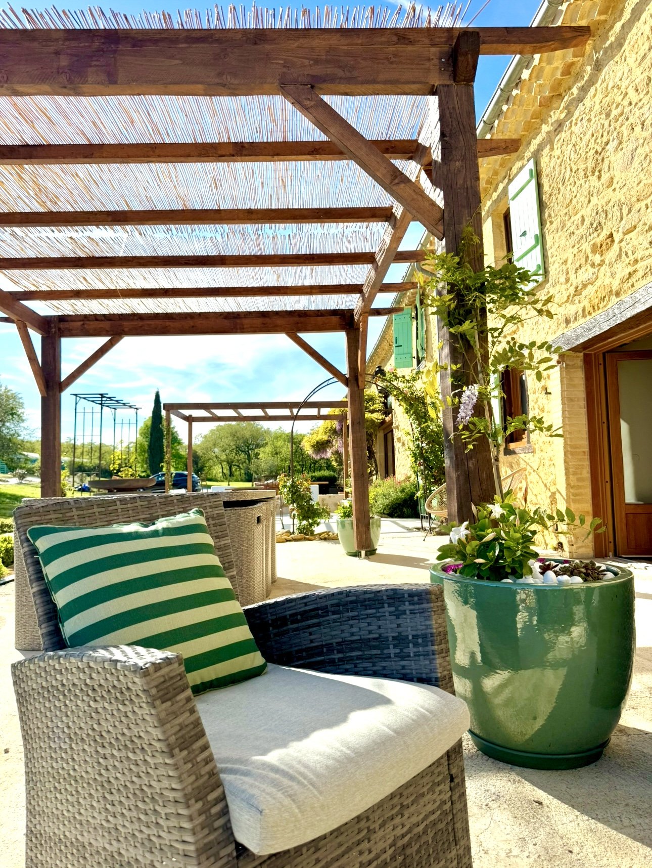 Une terrasse ensoleillée avec un fauteuil en rotin avec coussin rayé vert et blanc, une grande plante en pot vert, et une pergola en bois au-dessus, adjacente à une maison en pierre avec volets verts. En arrière-plan, un espace vert avec des arbres e