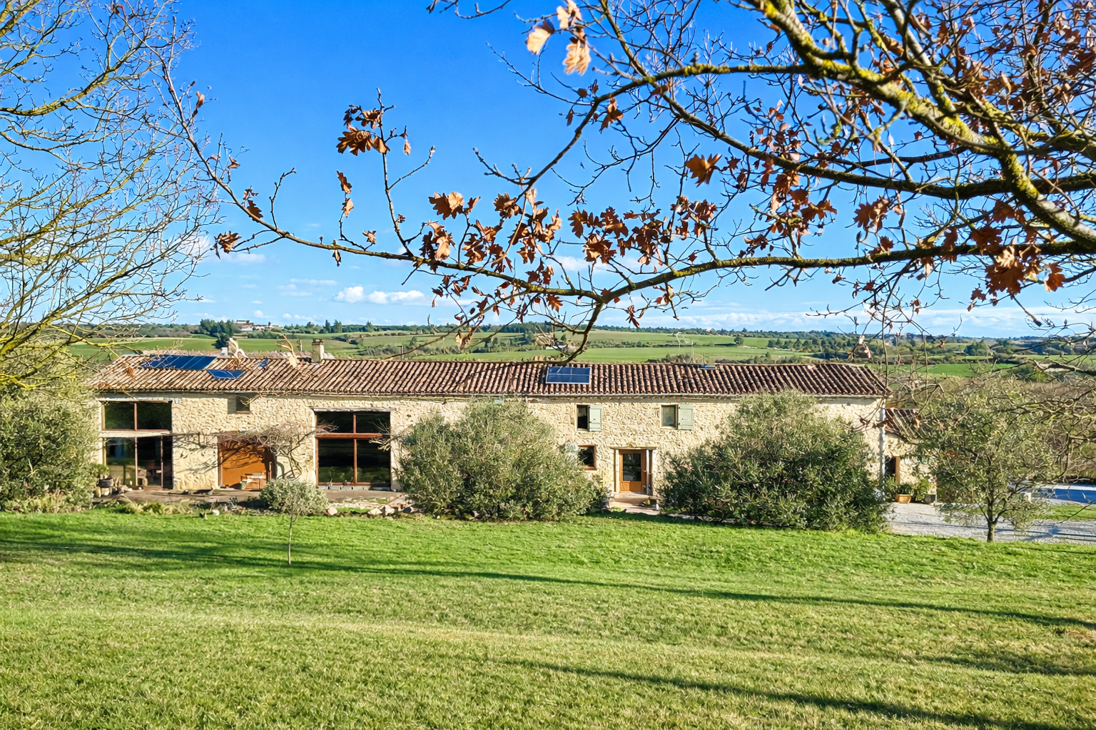 Une maison en pierre avec un toit en tuiles rouges, entourée d'arbres et de buissons, avec une pelouse verte devant, sous un ciel bleu avec quelques nuages.