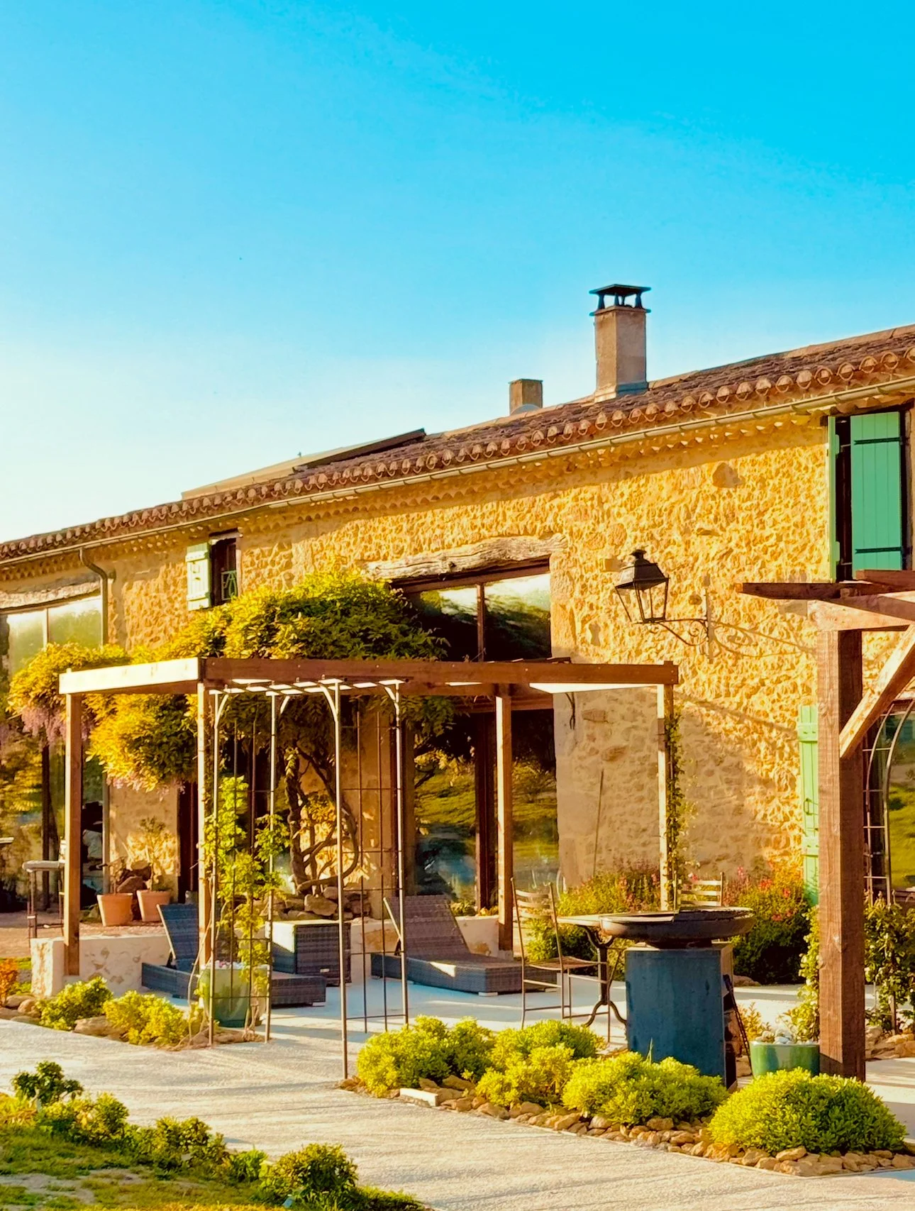 Maison en pierre avec un toit en tuiles, fenêtres vertes, et un espace extérieur avec mobilier, pergola et plantes. Ciel bleu au coucher du soleil.