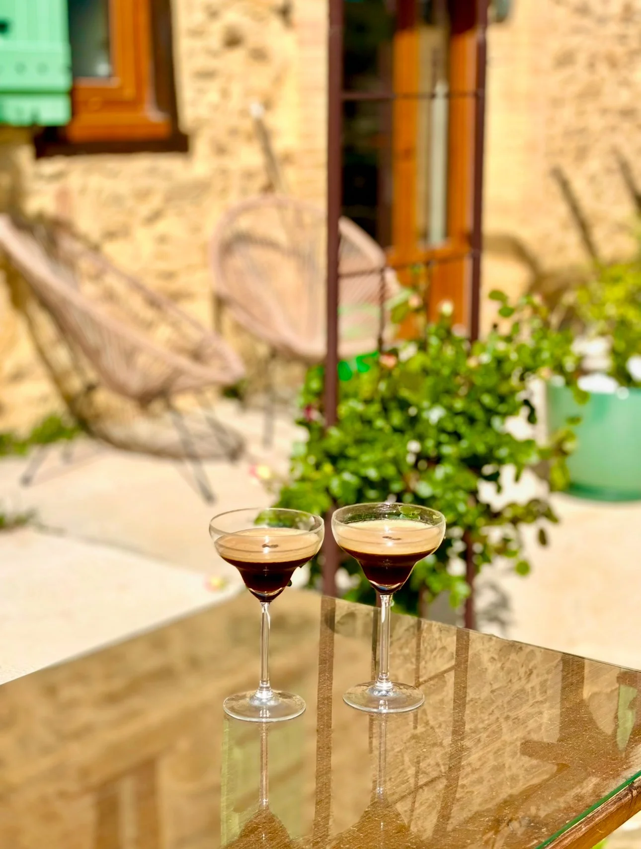 Deux coupes à champagne remplies de boisson rouge foncé sur une table en marbre, avec un décor de jardin en arrière-plan comprenant un mur en pierre, une chaise en rotin, une fenêtre en bois, des plantes vertes et un support pour plantes.