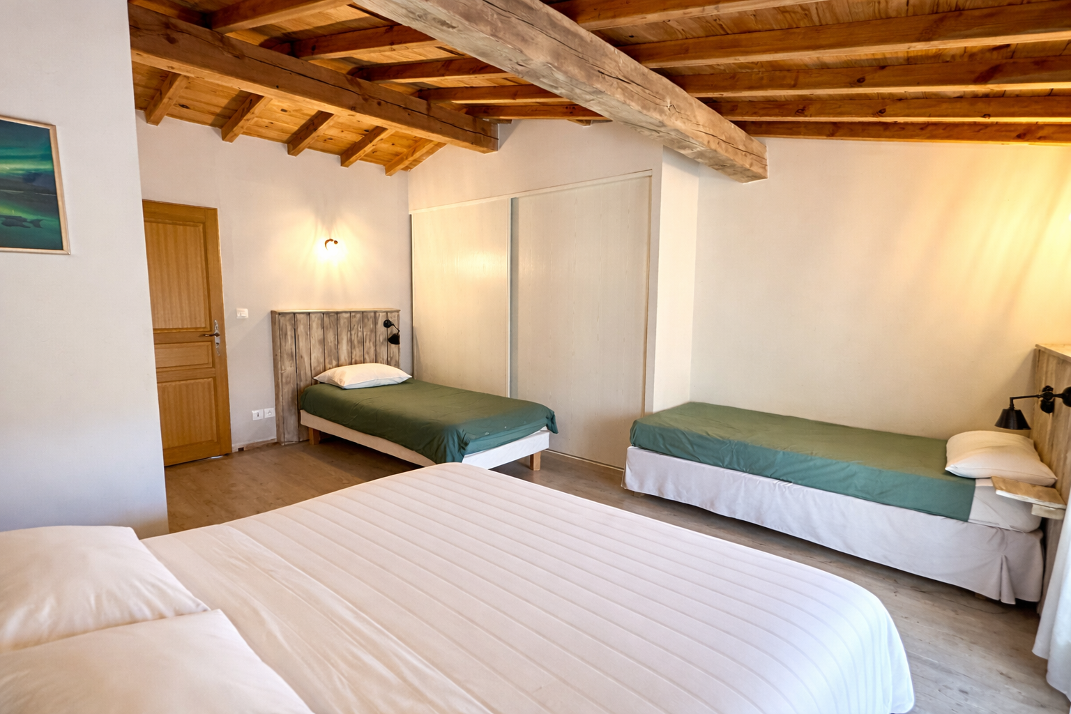 Chambre avec trois lits : un lit double au premier plan, deux lits simples avec tête en bois derrière, murs blancs, plafond en bois avec poutres apparentes, une peinture à l'huile sur le mur gauche, porte en bois, lampes murales noires.