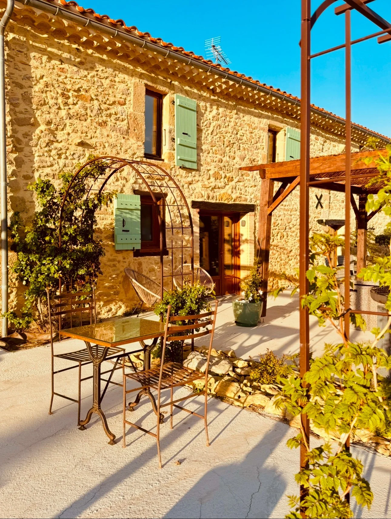 Maison en pierre avec volets verts, jardin avec mobilier en métal et pergola en bois, au soleil, ciel bleu.