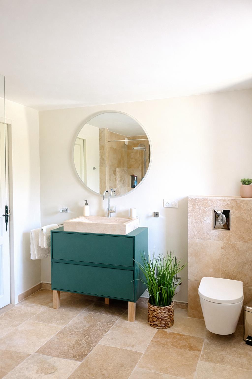 Salle de bains avec un mur blanc, un miroir rond, un lavabo en pierre sur une armoire verte, une plante en pot, un espace bain avec douche et une cuvette de toilettes