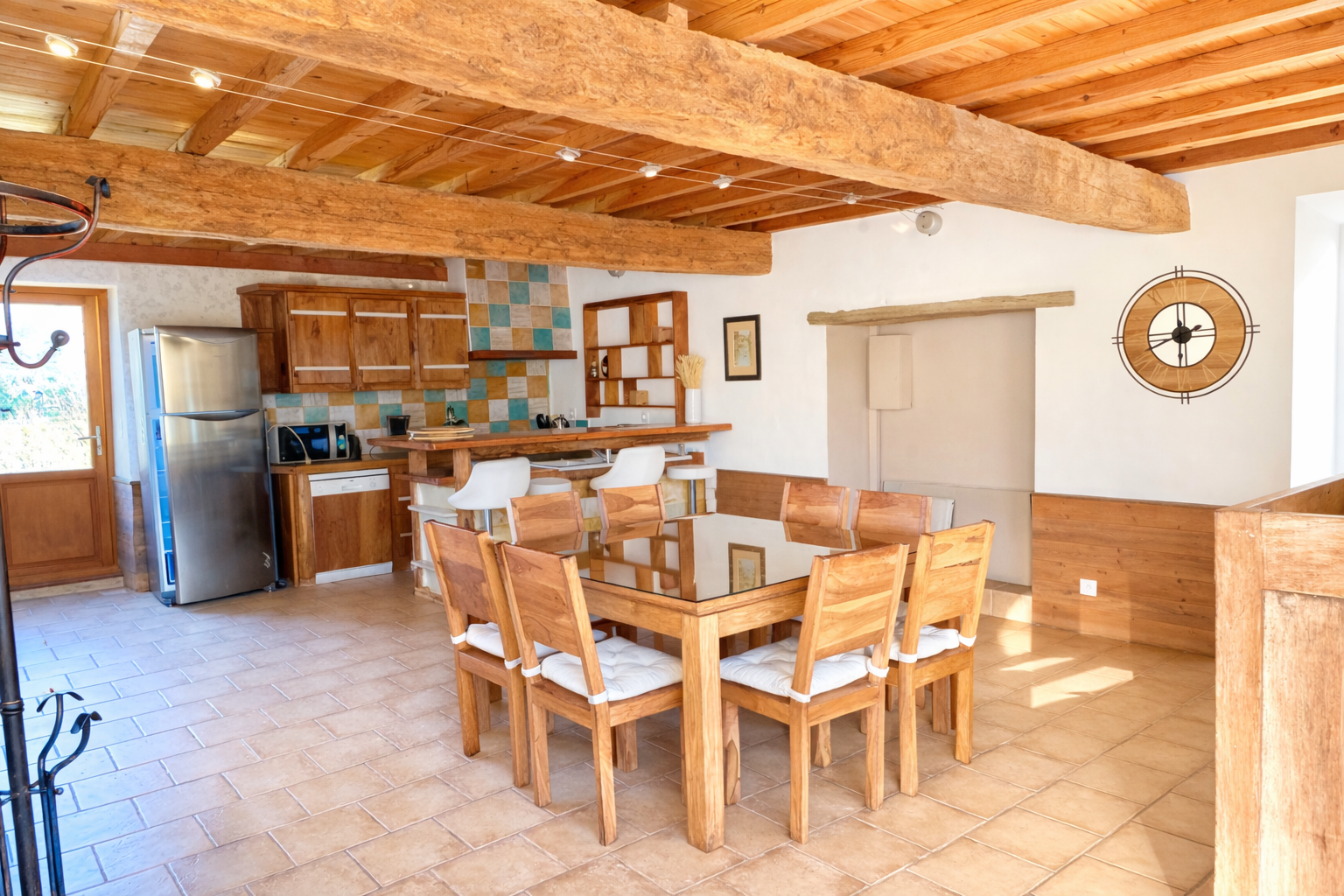 Intérieur d'une cuisine et salle à manger en bois, avec une table en bois et des chaises, un réfrigérateur en acier inoxydable, un comptoir avec quelques appareils électroménagers, un mur avec des carreaux colorés, et un mur blanc avec une horloge.