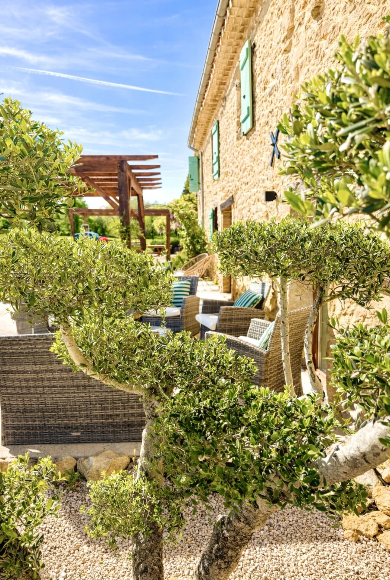 Une terrasse en pierre avec des sièges en rotin et des coussins rayés, entourée de buissons verts, face à une maison en pierre avec des volets verts, sous un ciel bleu avec quelques nuages.