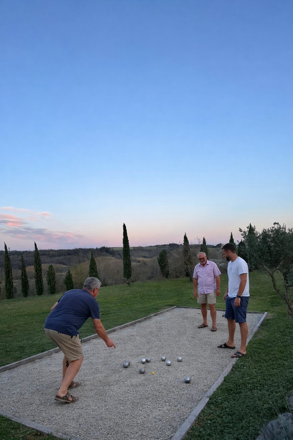 Trois hommes jouent à la pétanque sur une surface en gravier dans un paysage rural avec des arbres et des collines au coucher du soleil.