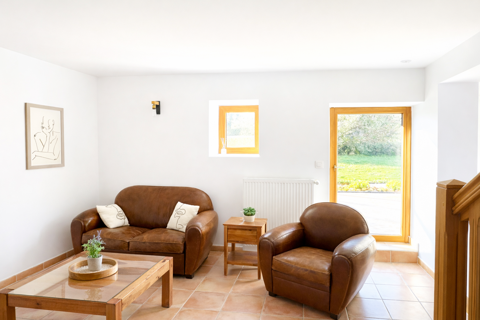 Salon avec deux fauteuils en cuir marron, table en bois avec un vase de fleurs, et deux coussins blancs avec des dessins. Mur blanc avec une œuvre d'art, deux petites fenêtres, et une porte-fenêtre donnant sur un jardin.