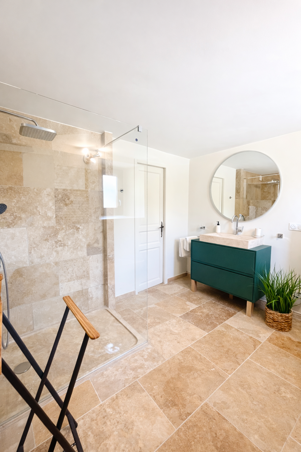 Salle de bain avec douche à l'italienne, meuble de rangement vert, miroir rond, lavabo en pierre, plante verte en pot, murs blancs et sol en carreaux beige, porte blanche, produits de toilette sur le lavabo.