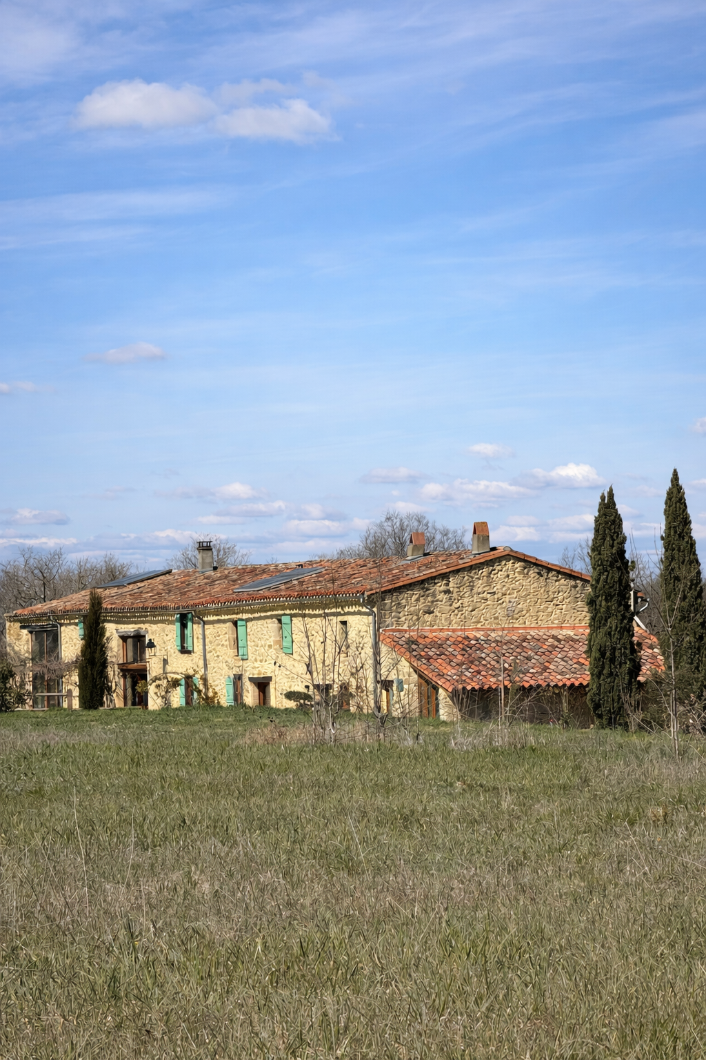 Une maison en pierre avec un toit en tuiles rouges, entourée d'arbres, sous un ciel bleu avec quelques nuages.