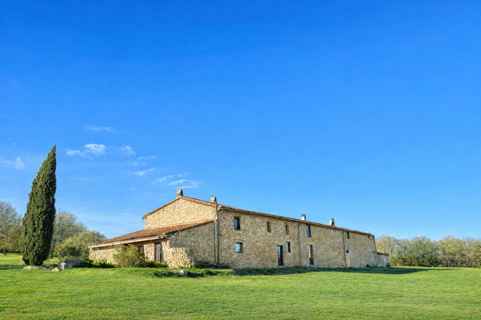 Maison en pierre au bord d'un grand espace vert sous un ciel bleu clair.
