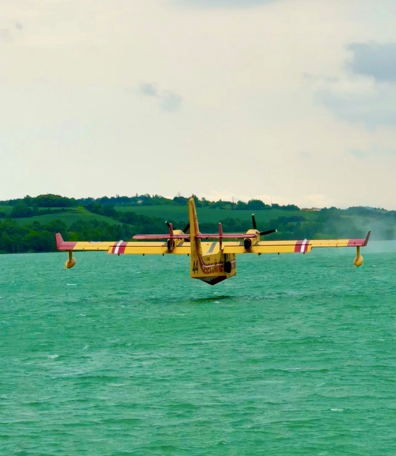 Un avion de pompiers survole une étendue d'eau verte, avec des collines verdoyantes en arrière-plan.