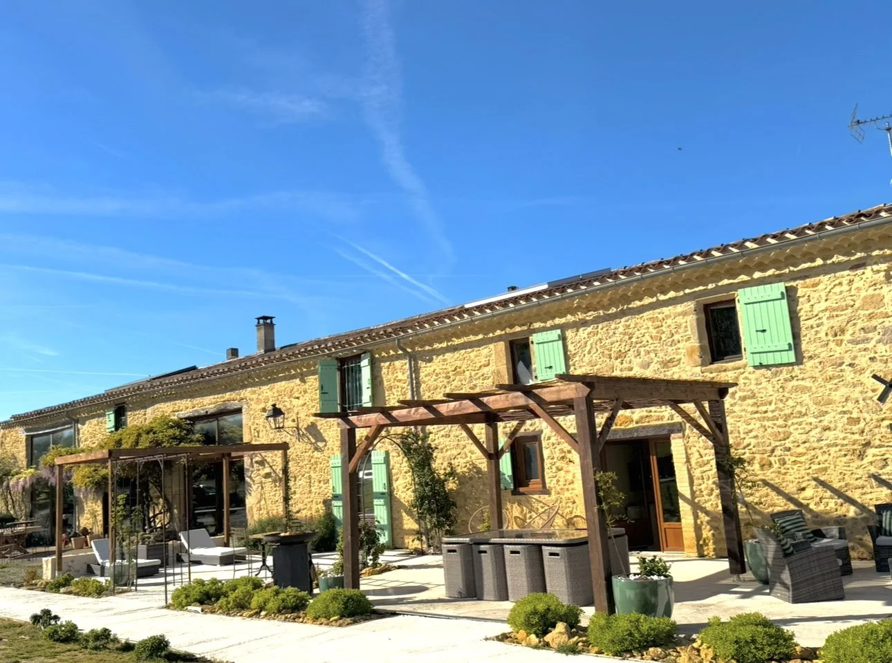 Maisons en pierre avec volets verts, terrasse extérieure avec mobilier, pergolas en bois, plantes et décorations extérieures, ciel bleu clair.