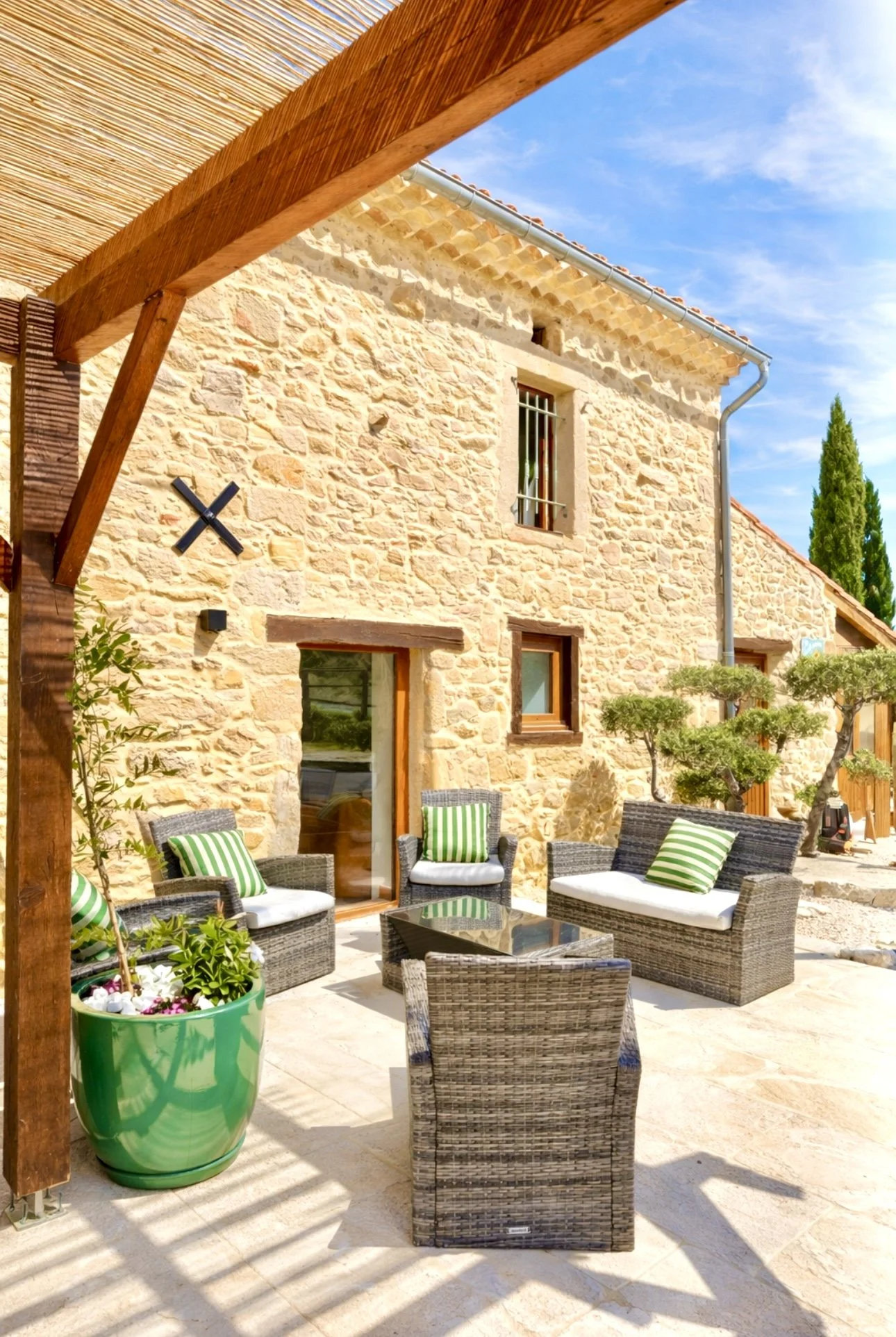 Espace extérieur avec mobilier en rotin, plusieurs fauteuils avec coussins à rayures vertes et blanches, table en verre, plantes en pot vert, maison en pierre avec fenêtres, ciel bleu, arbres en arrière-plan.