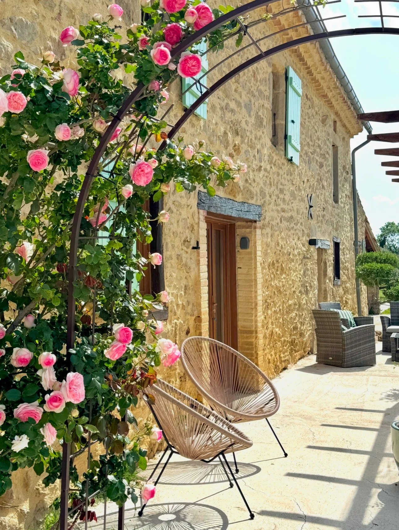 Une maison en pierre beige avec volets bleus, entourée de fleurs roses et d'un mobilier d'extérieur moderne, sous un ciel ensoleillé.